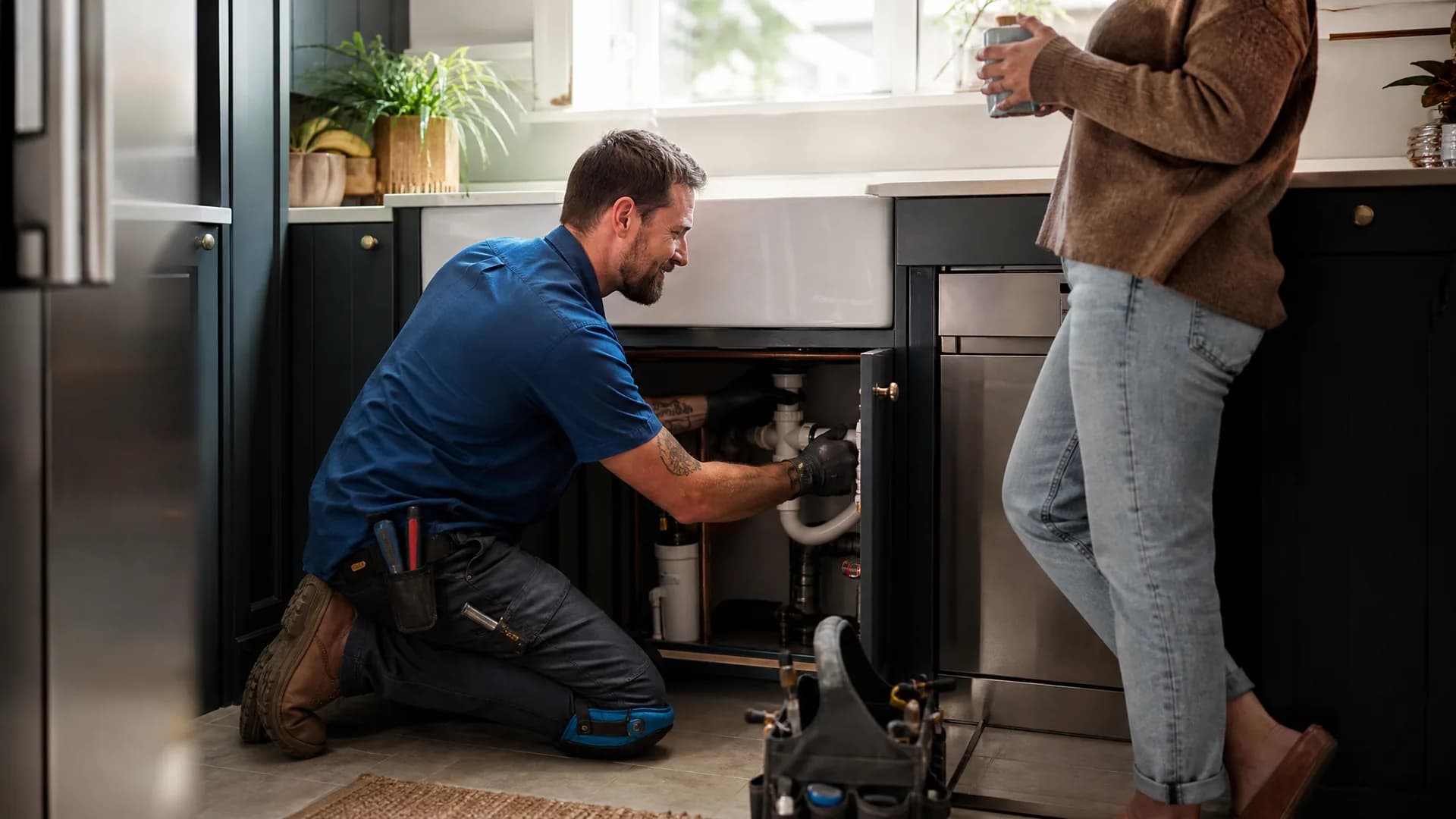 A plumber fixing pipework under a kitchen sink while the homeowner stands nearby with a mug